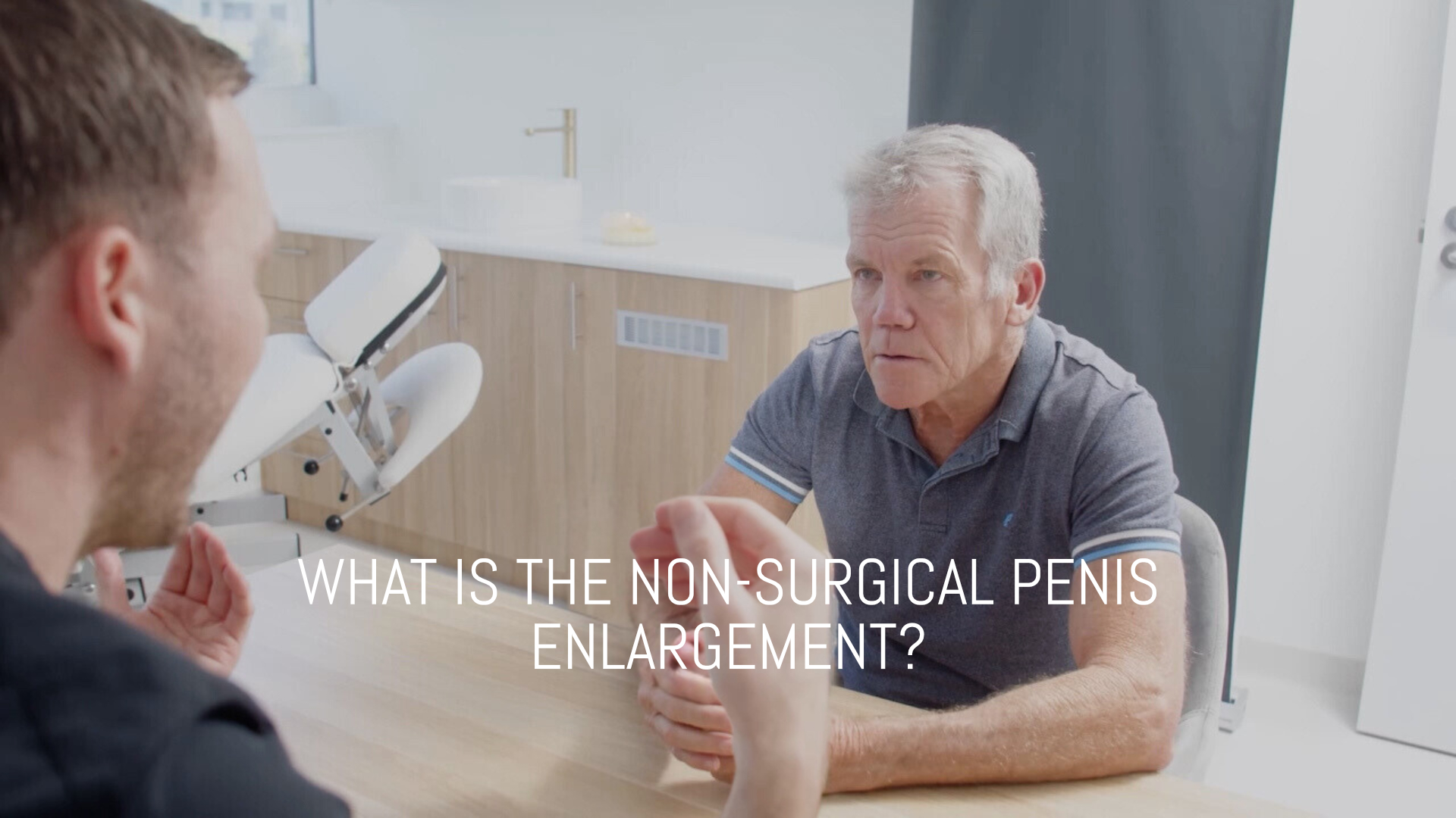 A man with white hair sits at a table, listening intently to another person facing him. The text on the image reads, What is the non-surgical penis enlargement? In the background, theres a dental or medical office setup.