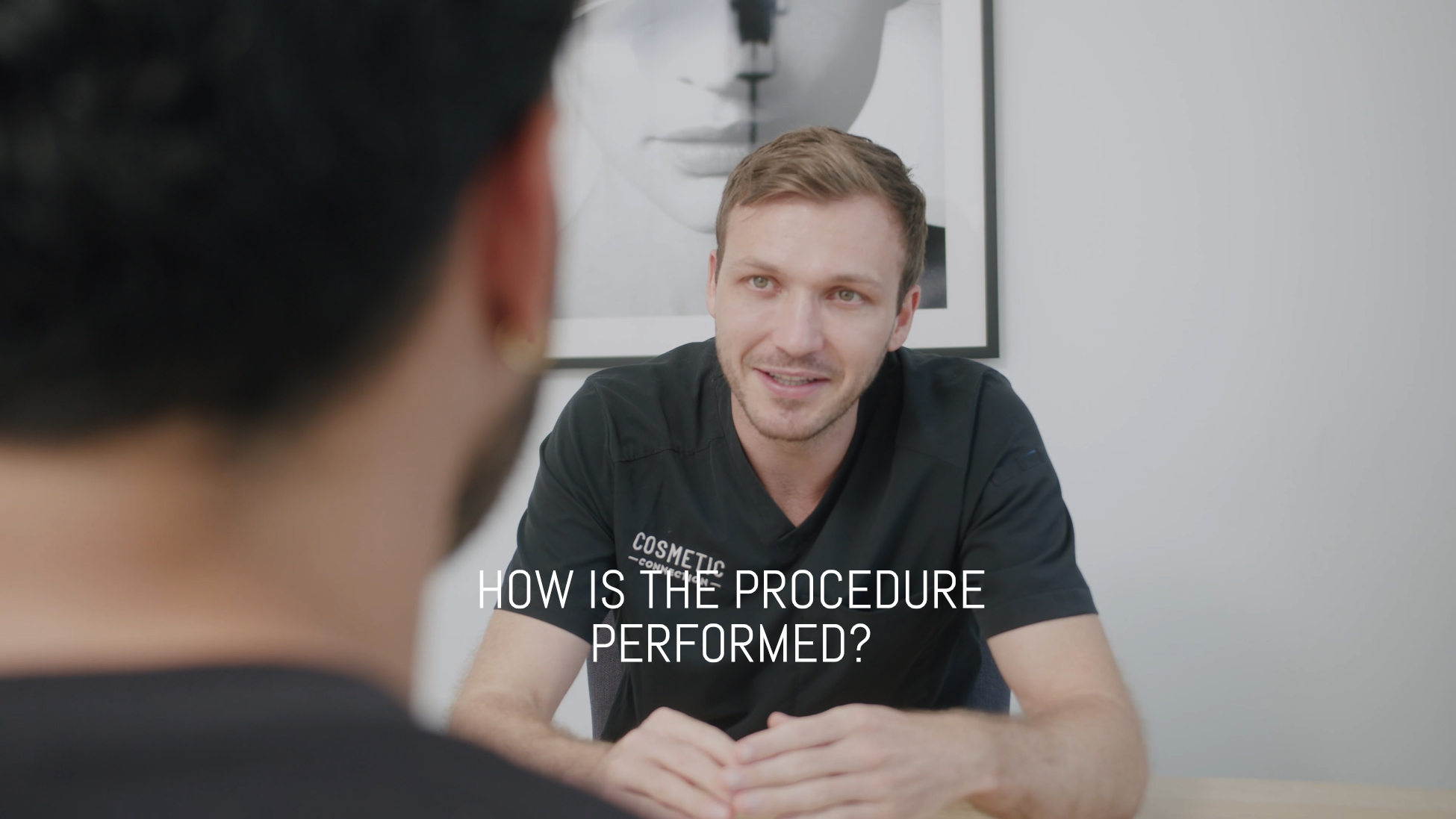 A man in a black shirt sits at a desk speaking to another person, whose back is to the camera. The text How is the procedure performed? is overlaid. A black-and-white portrait hangs in the background.