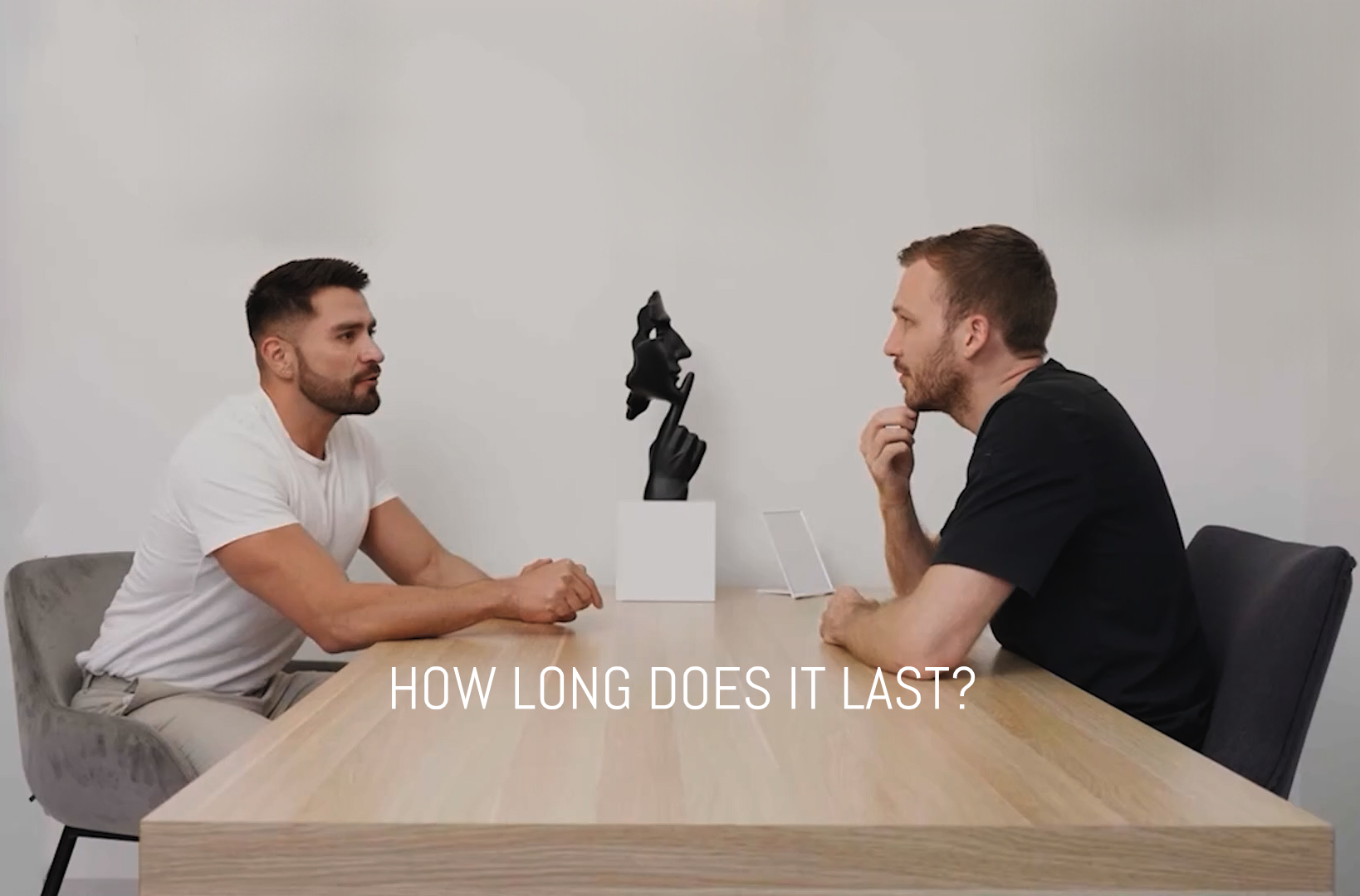 Two men sit across from each other at a wooden table with a small black sculpture between them. Both are engaged in conversation. The text HOW LONG DOES IT LAST? is overlaid at the bottom of the image.