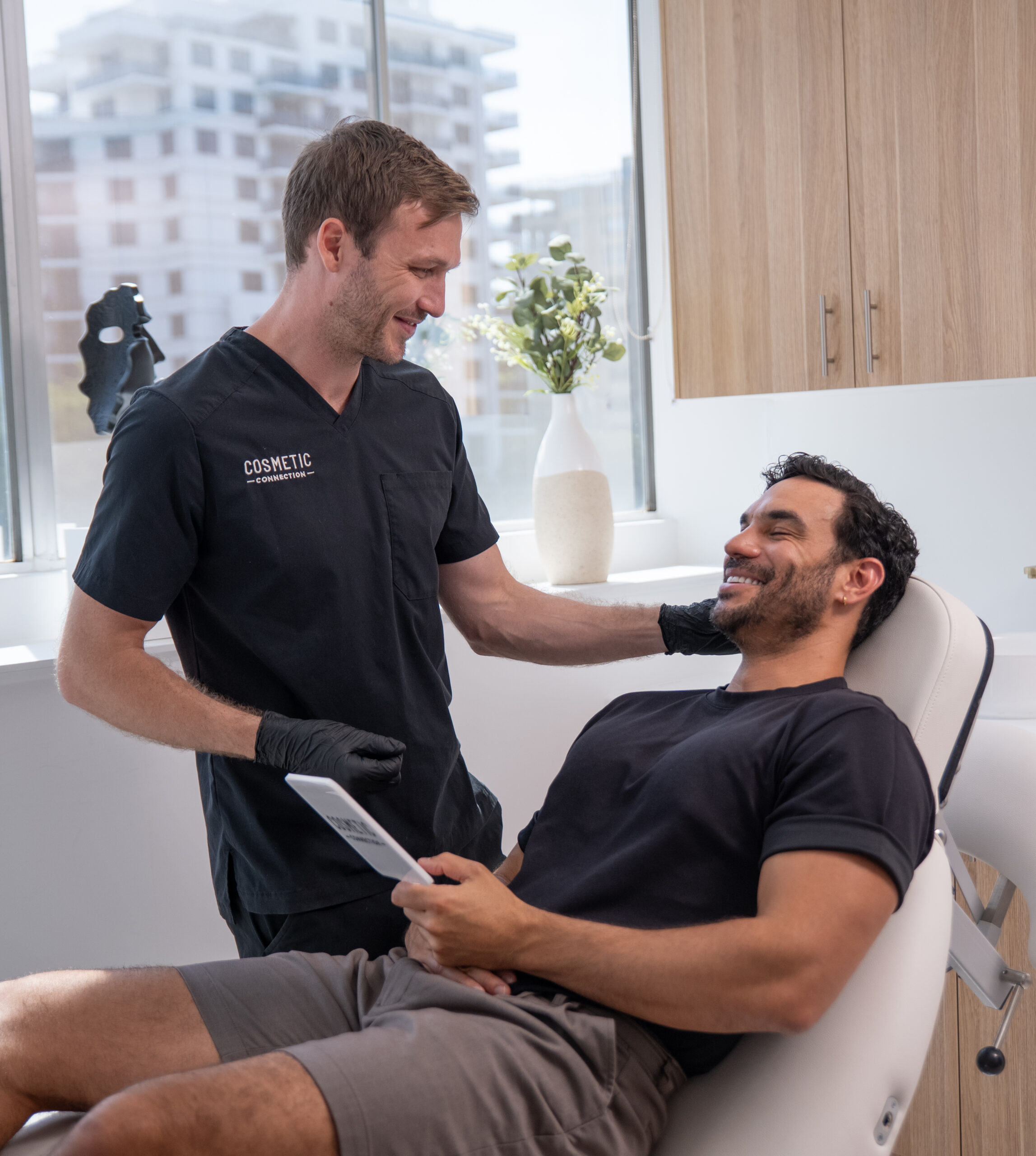 A man in a dental or medical setting, wearing black scrubs, smiles and talks to a seated patient holding a tablet. The setting appears bright and modern, with a large window and decorative items in the background.