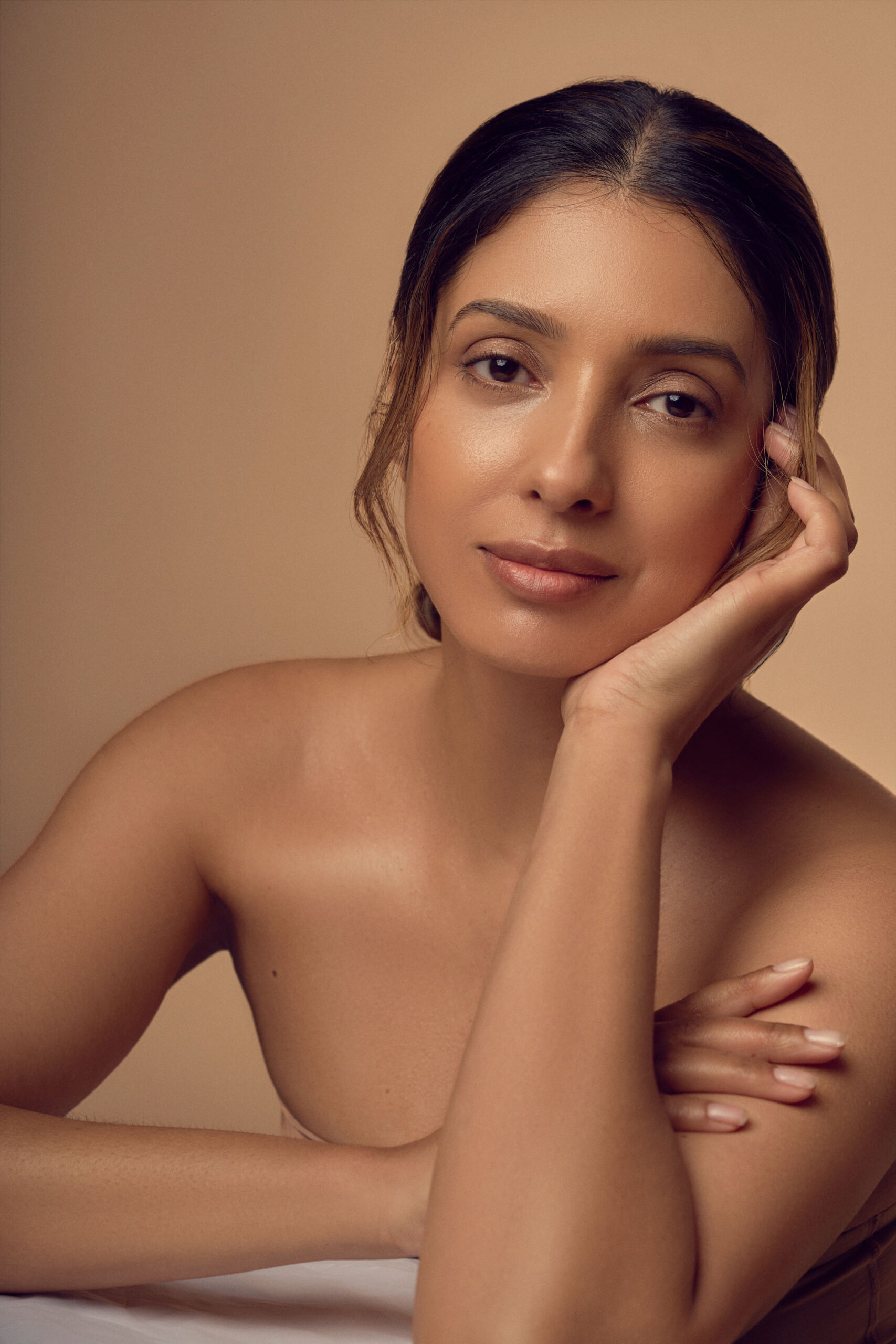 A woman with long, dark hair pulled back, resting her chin on her hand. She has a calm expression and is posed against a soft beige background, showcasing a natural look with minimal makeup.
