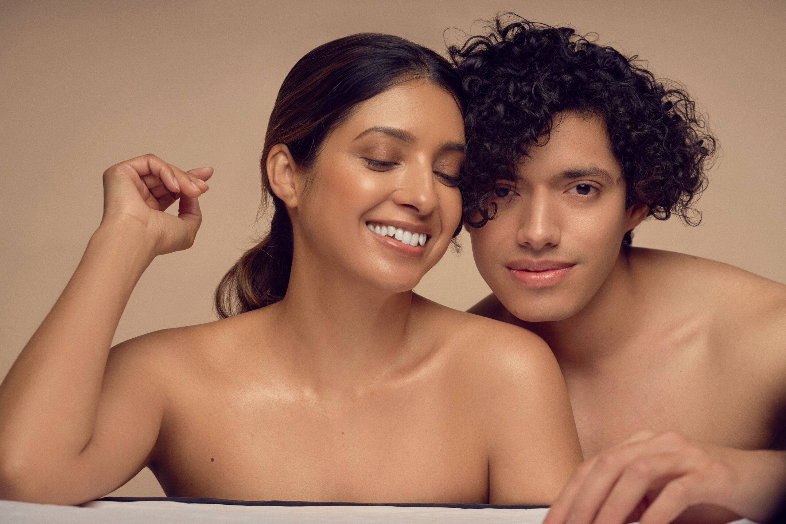 A couple with curly and wavy hair sits closely together against a neutral background. They are smiling and appear relaxed, with one person resting their head on the others shoulder.