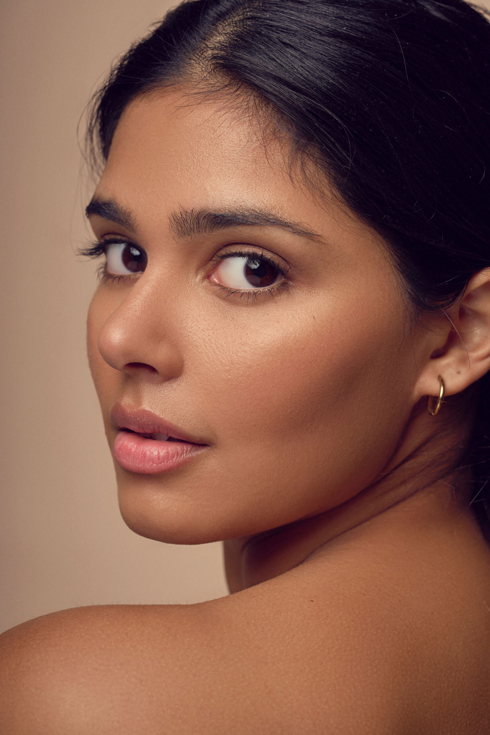 A woman with long dark hair is looking over her shoulder at the camera. She has subtle makeup and is wearing a small hoop earring. The background is a soft beige tone, highlighting her features.