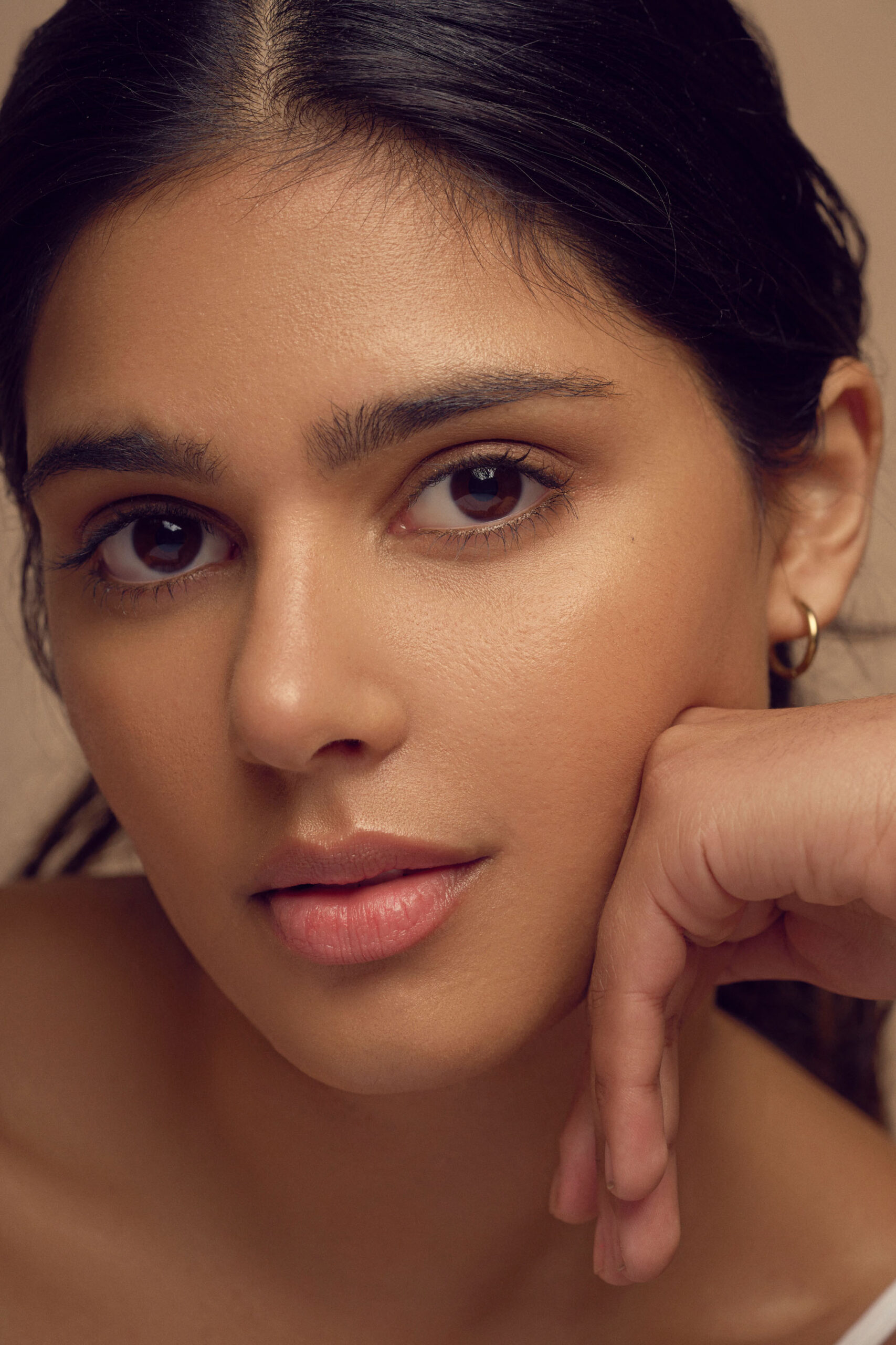A close-up of a person with long dark hair gazing into the camera, resting their chin on their hand. They have a calm expression, minimal makeup, and are wearing a small hoop earring. The background is neutral.