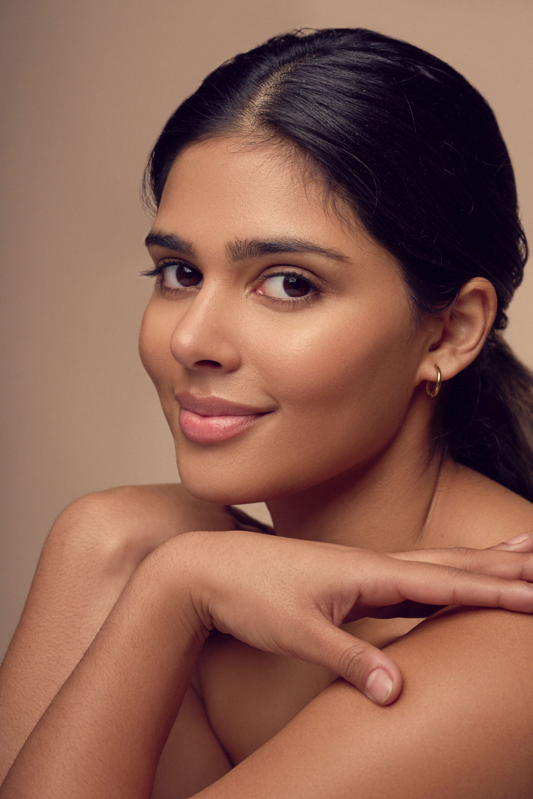 A woman with long dark hair pulled back, smiling gently. She rests her chin on her crossed arms. She has a warm skin tone and is wearing natural makeup. The background is a soft beige, creating a calm and elegant portrait.