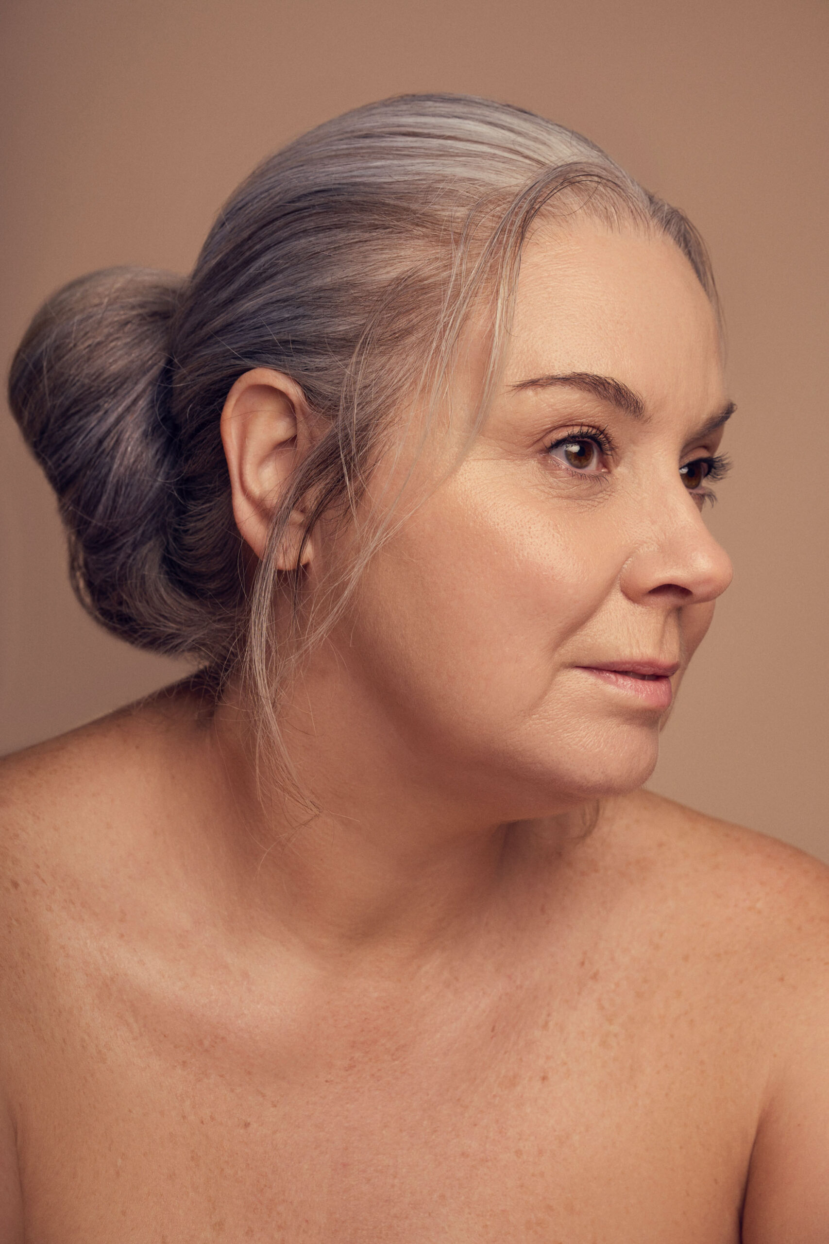 A woman with gray hair styled in a bun looks to the side against a neutral background. She appears thoughtful and calm, with a bare shoulder visible in the forefront.