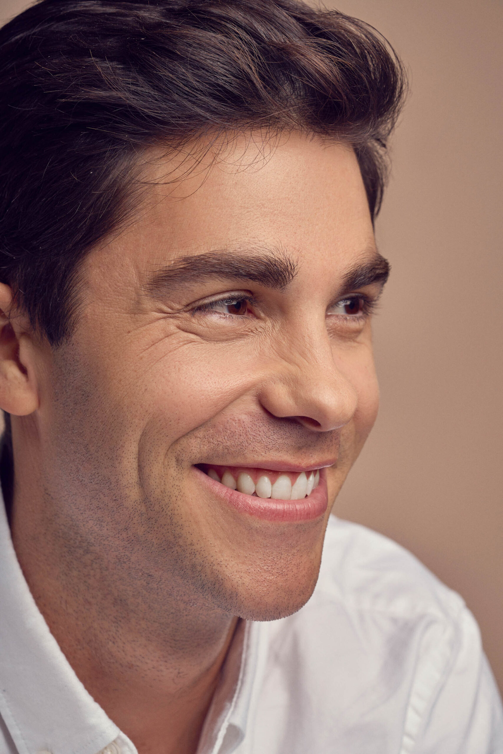 A man with dark hair is smiling broadly. He is wearing a white collared shirt, with a soft beige background. His expression is cheerful and relaxed.
