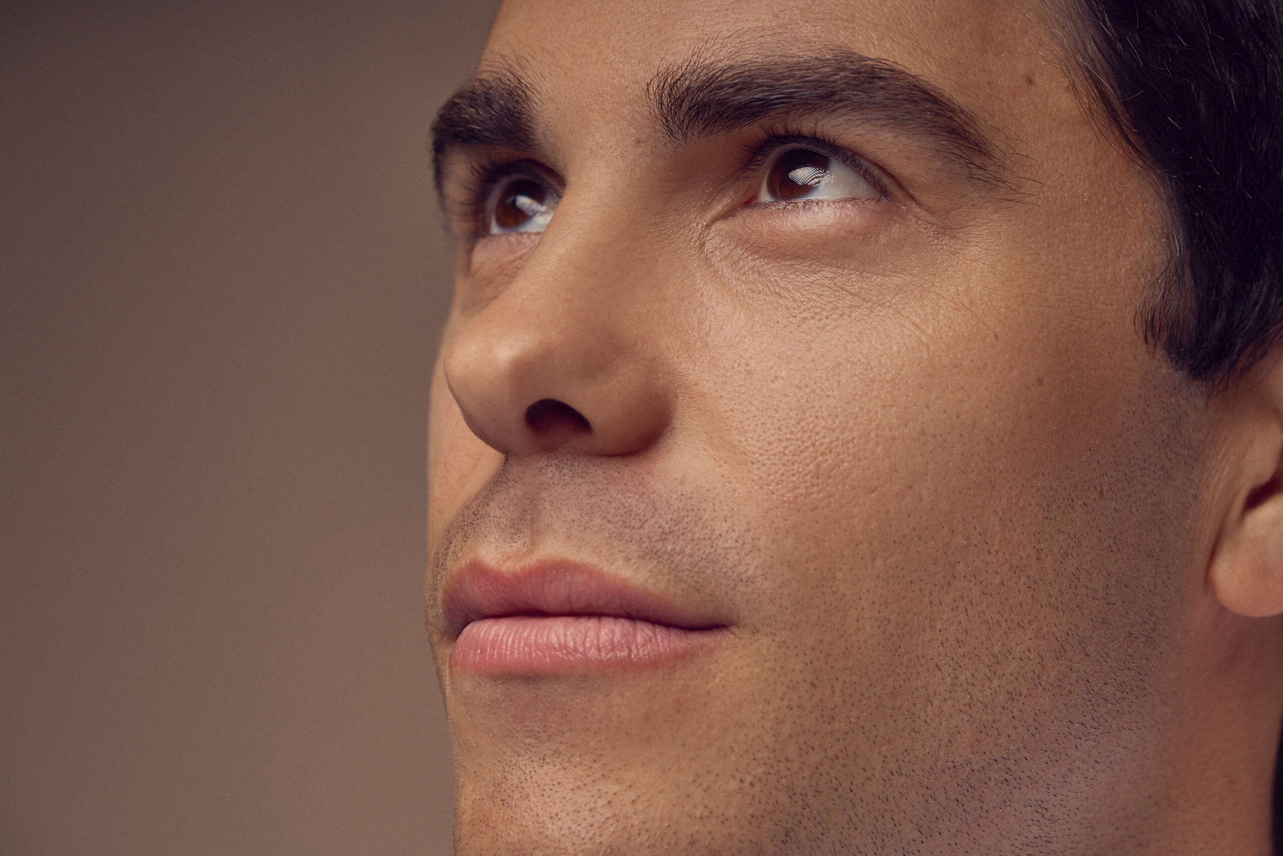 Close-up of a persons face looking upward with a neutral expression. The skin is smooth, eyebrows are dark and eyes are gazing skyward. The background is softly blurred, emphasizing the contemplative pose of the individual.