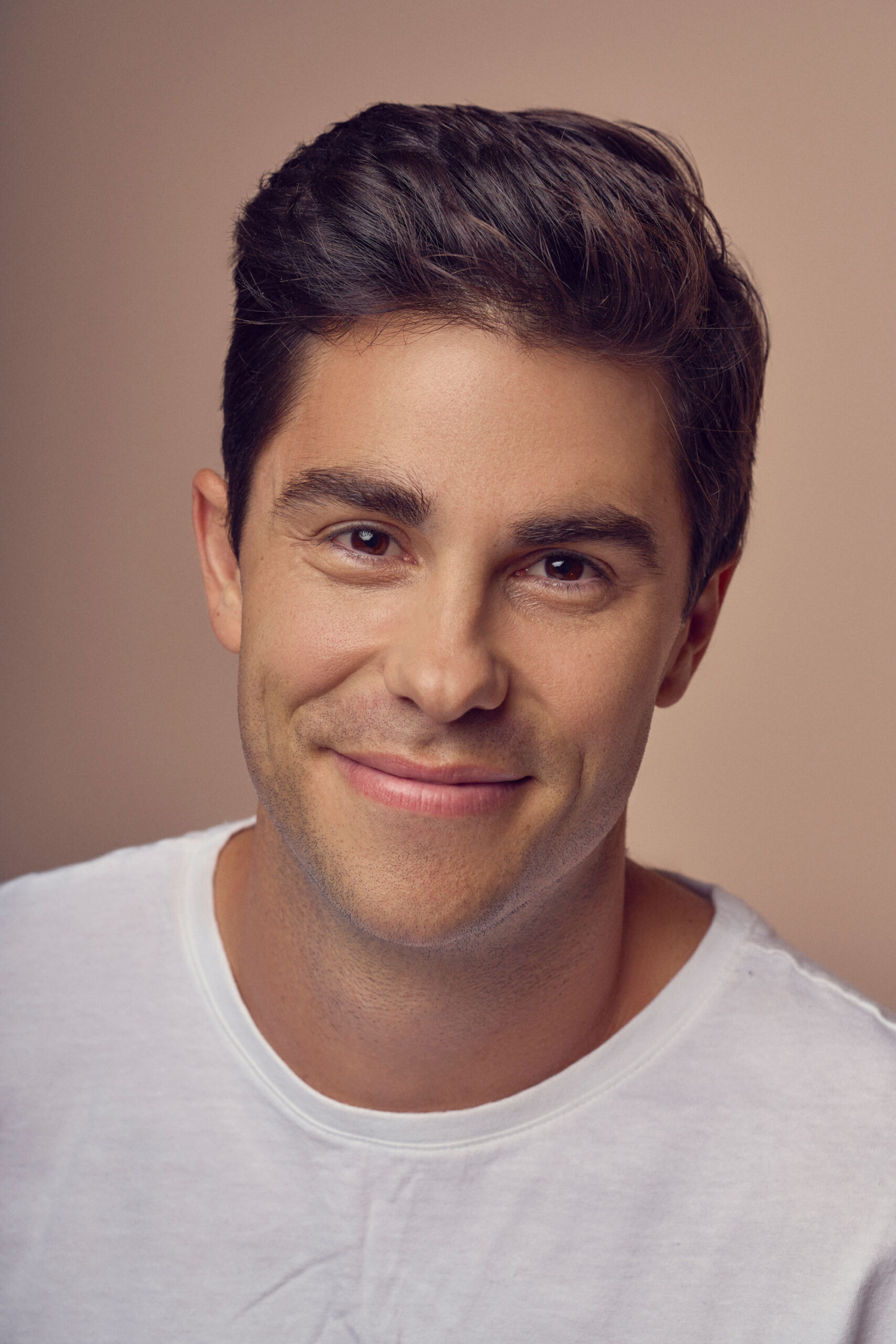 A person with short dark hair smiling at the camera, wearing a white shirt against a neutral background.