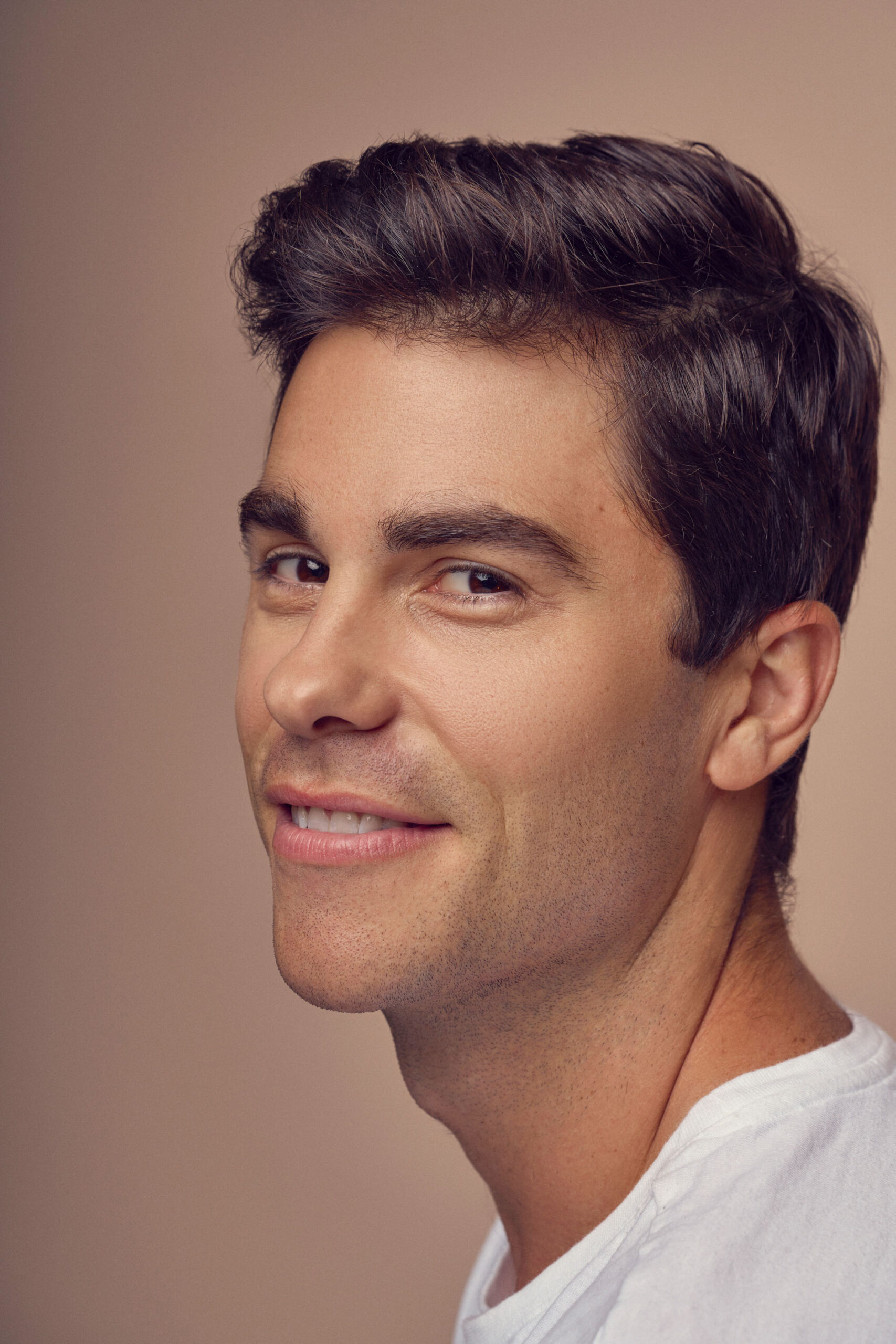 A man with short brown hair and a light complexion is smiling softly. He is wearing a white shirt, and the background is a neutral, warm tone.