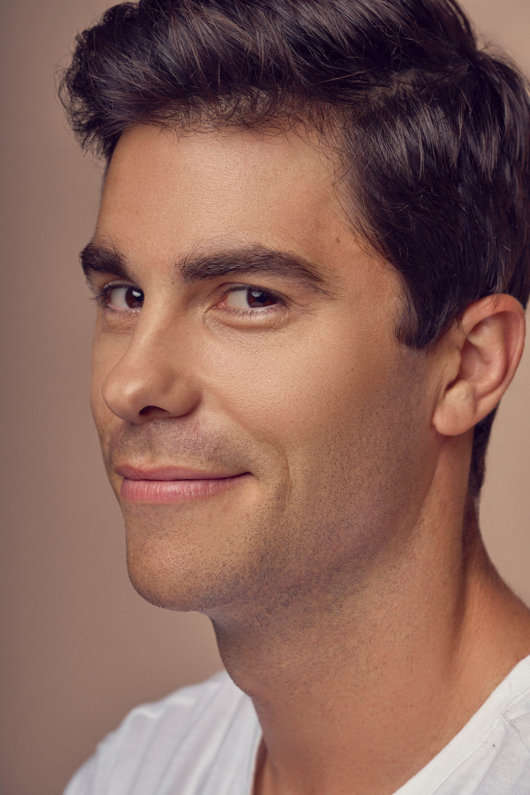 A person with short dark hair and a slight smile looks towards the camera. They are wearing a white shirt against a neutral background.