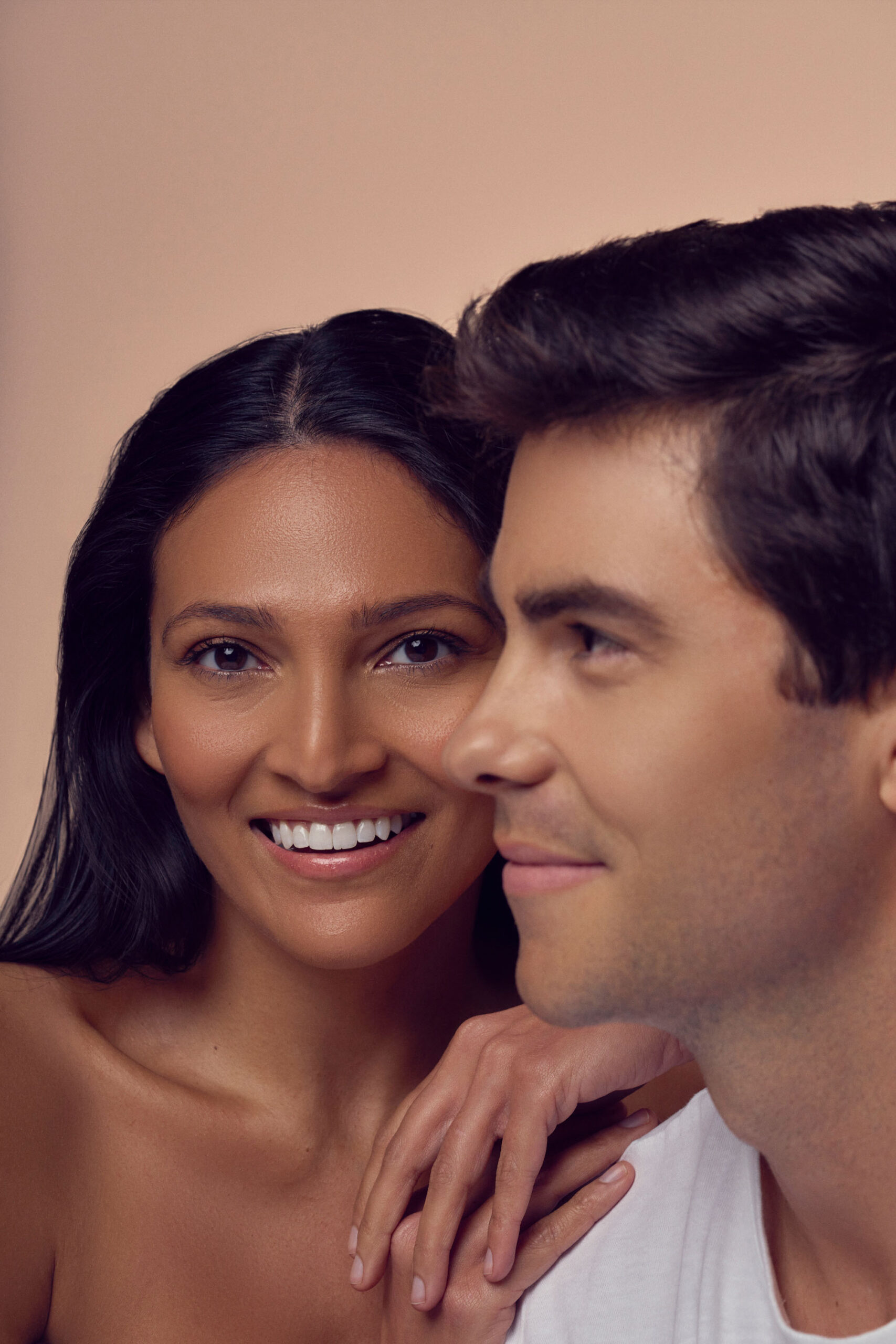 A woman with long dark hair smiles, resting her hand on the shoulder of a man beside her. The man, with short dark hair, is looking forward, wearing a white shirt. The background is a soft beige color.