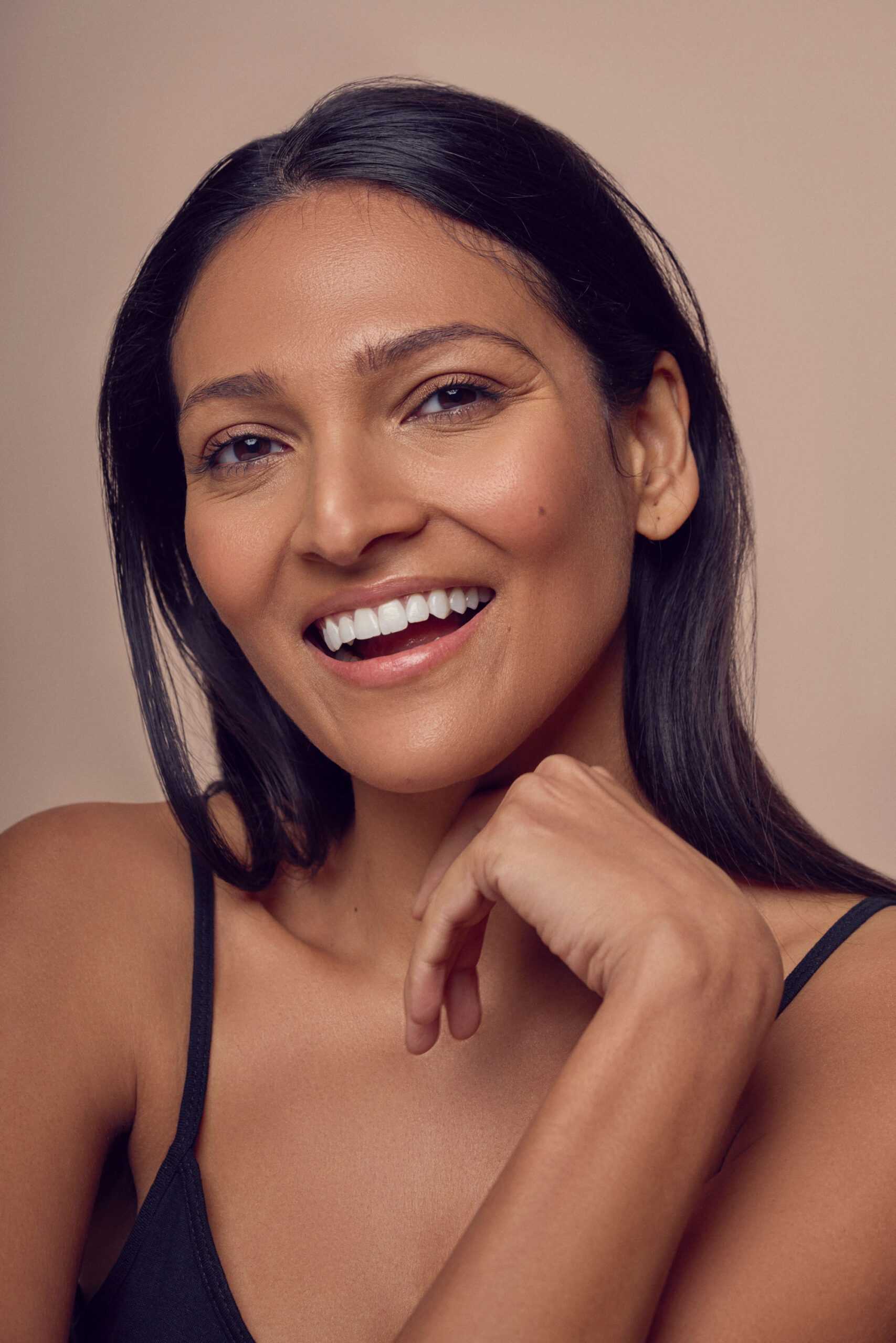 A person with long dark hair is smiling at the camera. They have one hand gently touching their chin. The background is a soft beige, and the person is wearing a sleeveless black top.