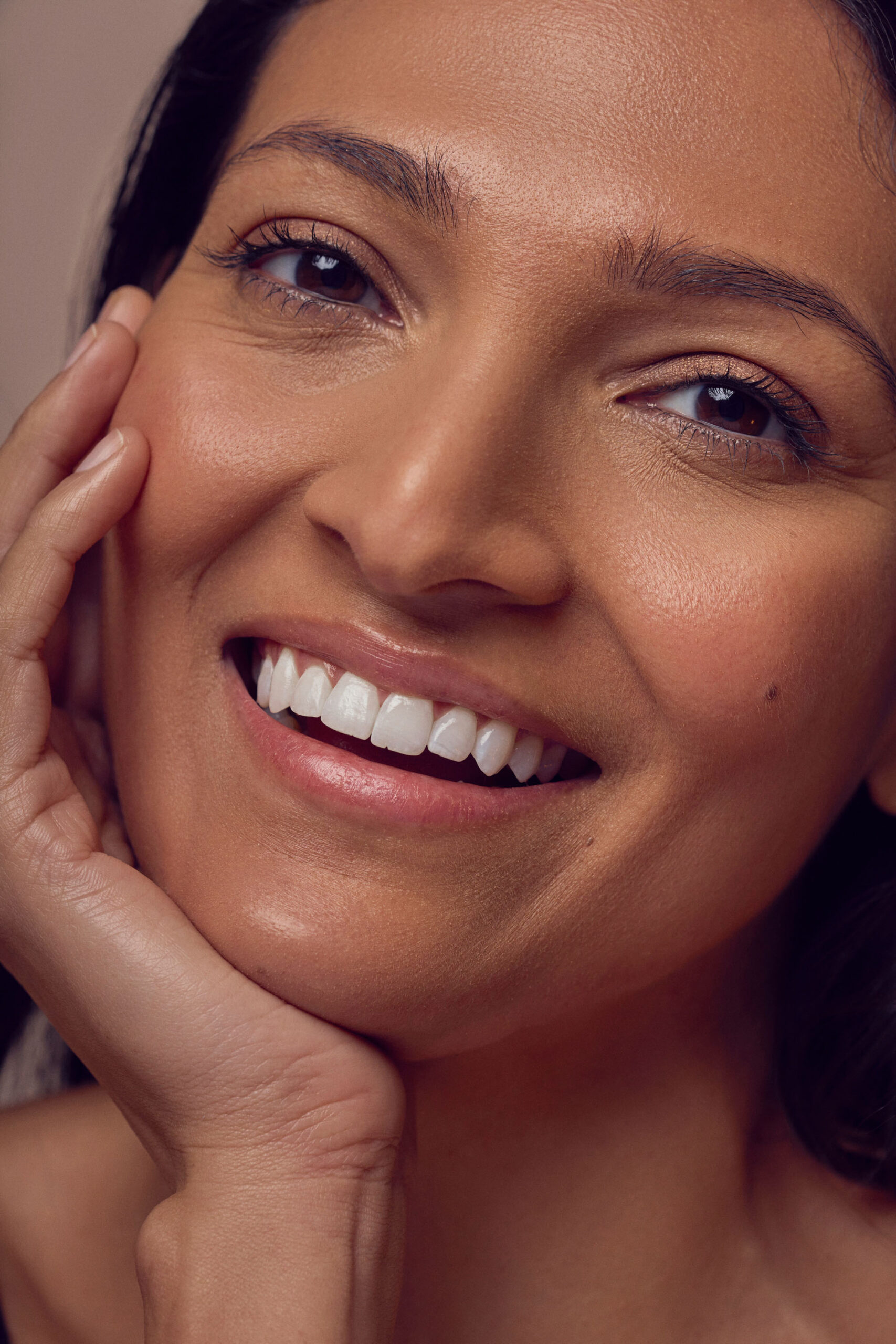 A person smiling with their hand gently resting on their cheek, showing bright white teeth and smooth skin.