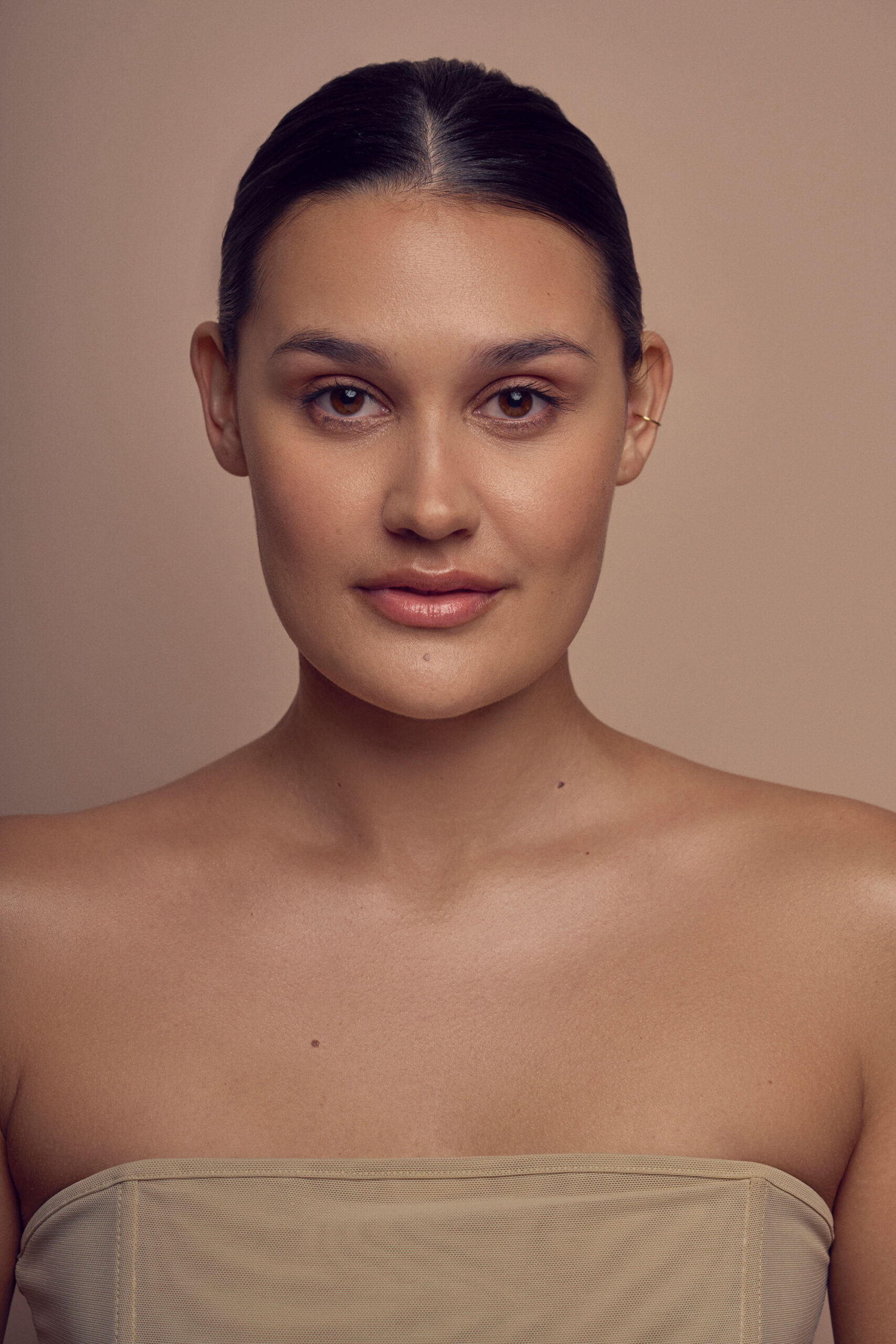 A person with dark hair pulled back, wearing a strapless beige top, is posing against a plain beige background. They have a neutral expression and are looking directly at the camera.