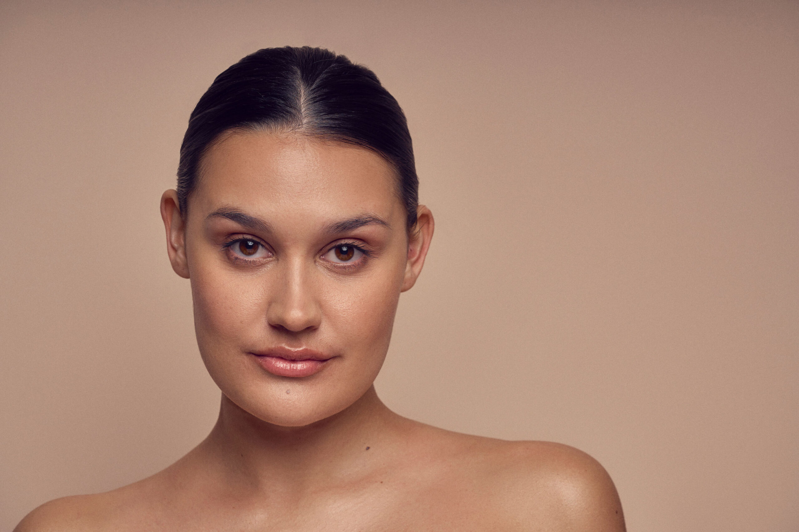 A woman with dark hair pulled back gazes directly at the camera against a neutral beige background. She has a calm expression and wears natural makeup, highlighting her features. Her shoulders are bare.