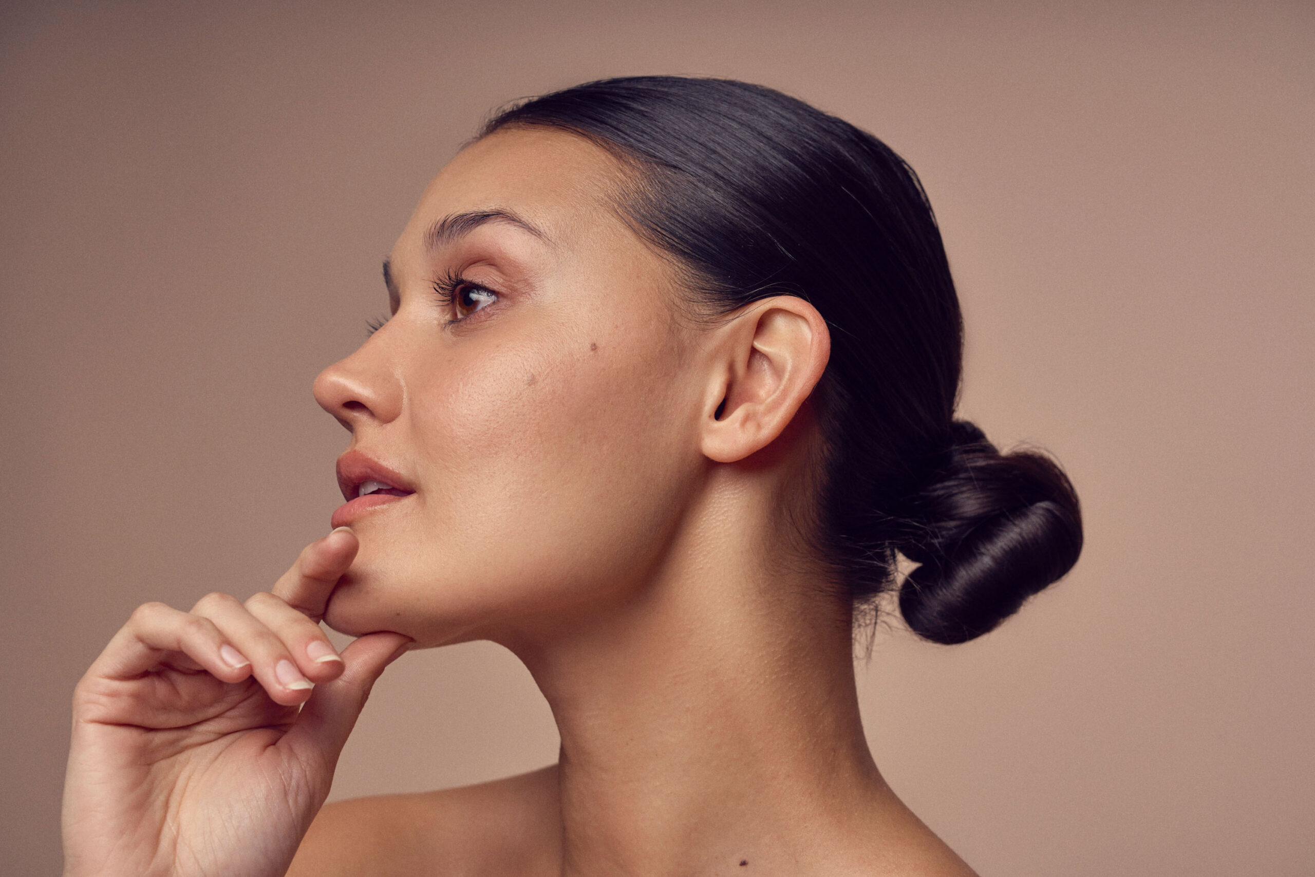 A woman with dark hair styled in a sleek bun is shown in profile against a neutral background. She rests her chin on her hand, appearing contemplative. Her skin is clear and her expression is serene.