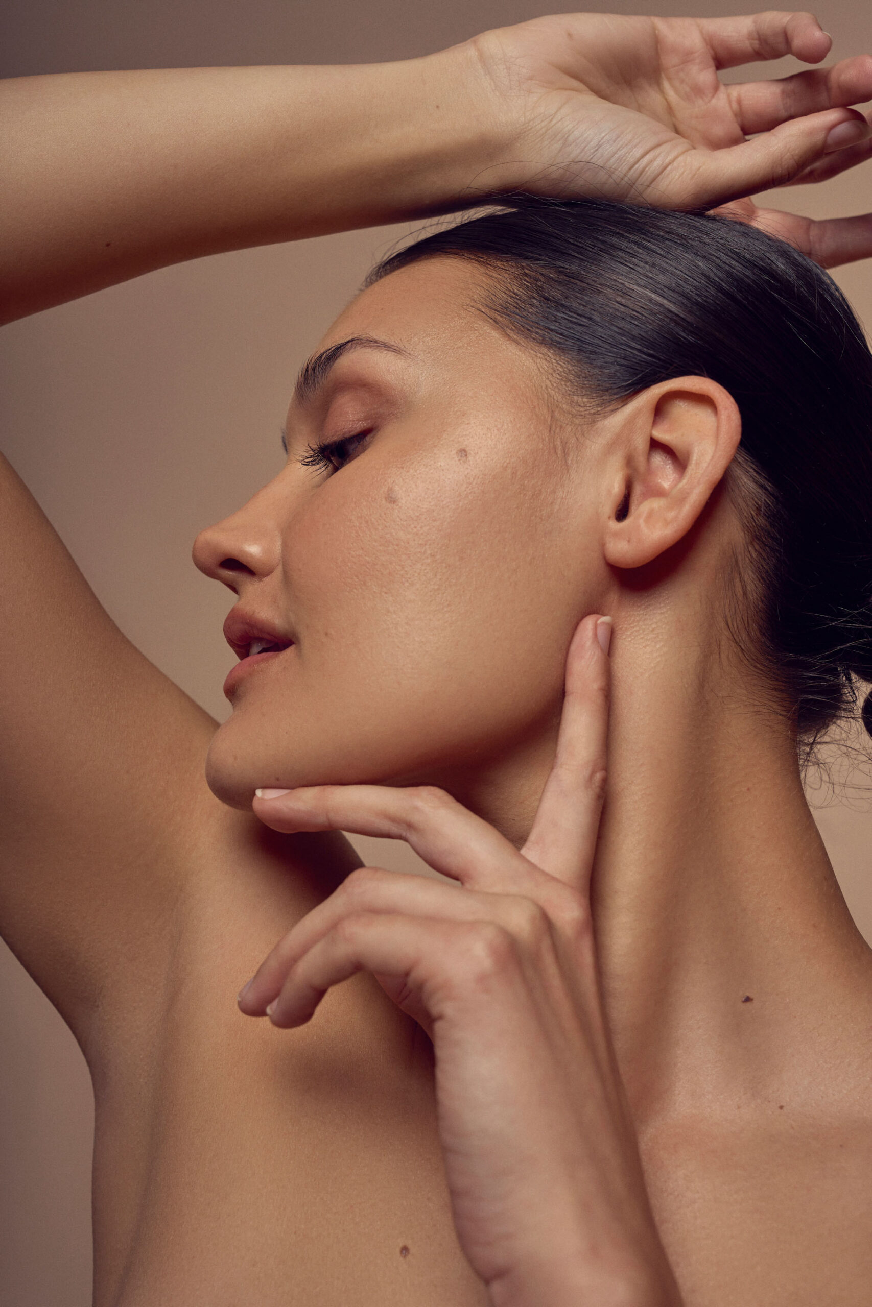 A woman with a smooth complexion poses against a neutral background. She has her hair pulled back and raises one arm over her head while gently touching her chin with the other hand, emphasizing her profile.
