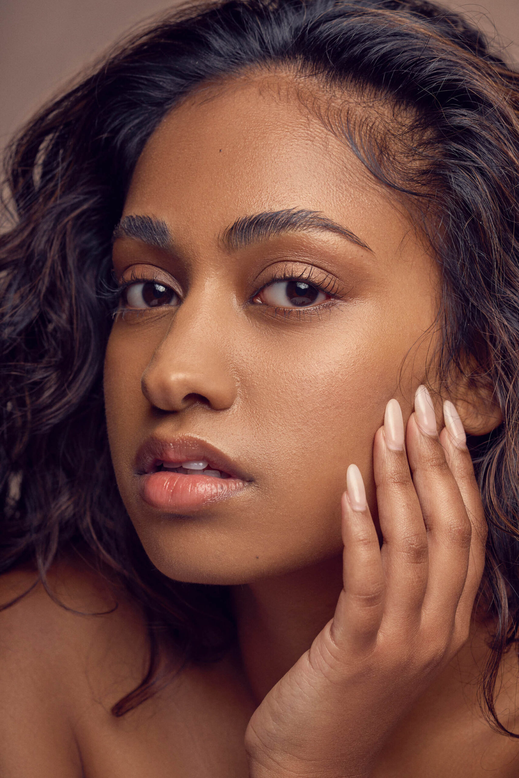 A close-up portrait of a woman with long, wavy hair and natural makeup. She rests her hand on her cheek, looking directly at the camera with a neutral expression. The background is a soft, muted tone.