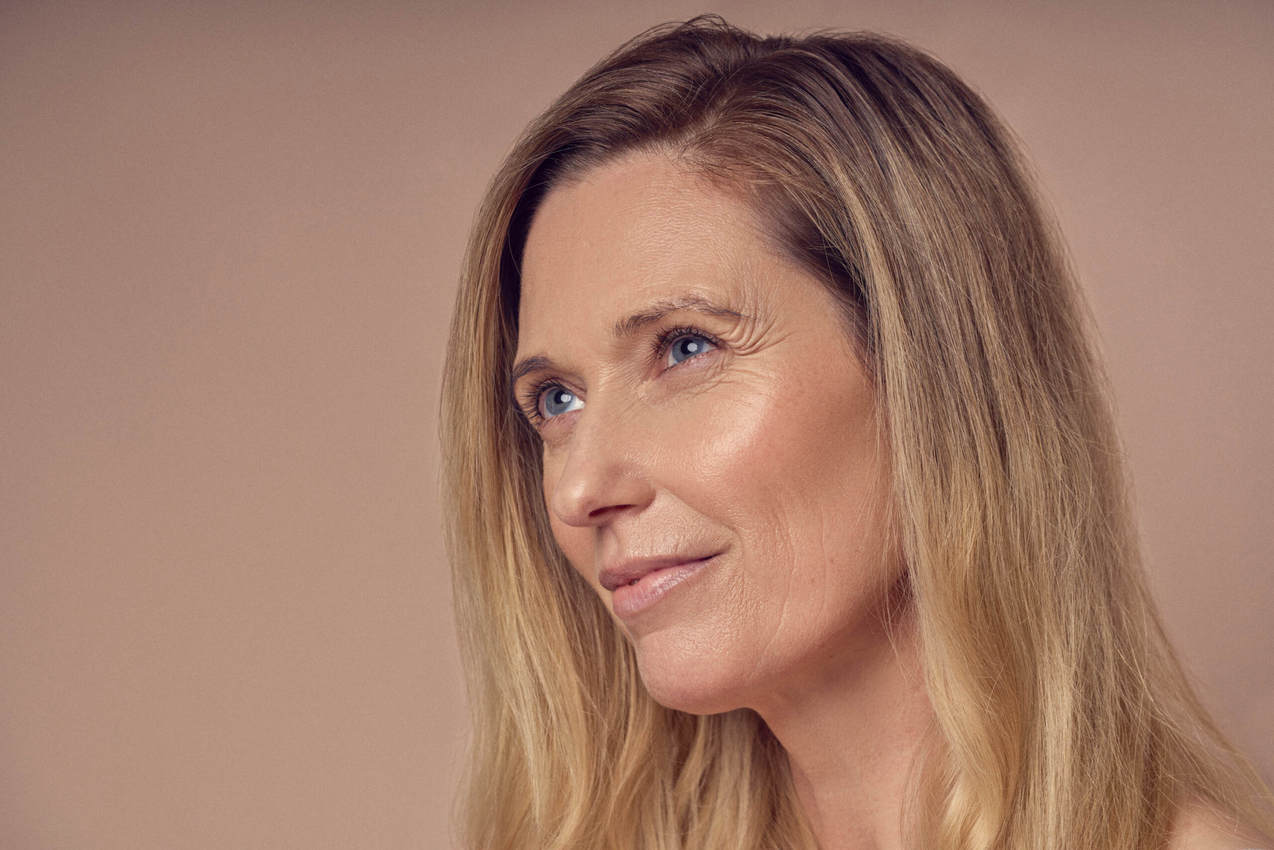 A woman with long blonde hair looks thoughtfully upwards against a neutral background. Her expression is serene, highlighting her blue eyes and subtle smile.