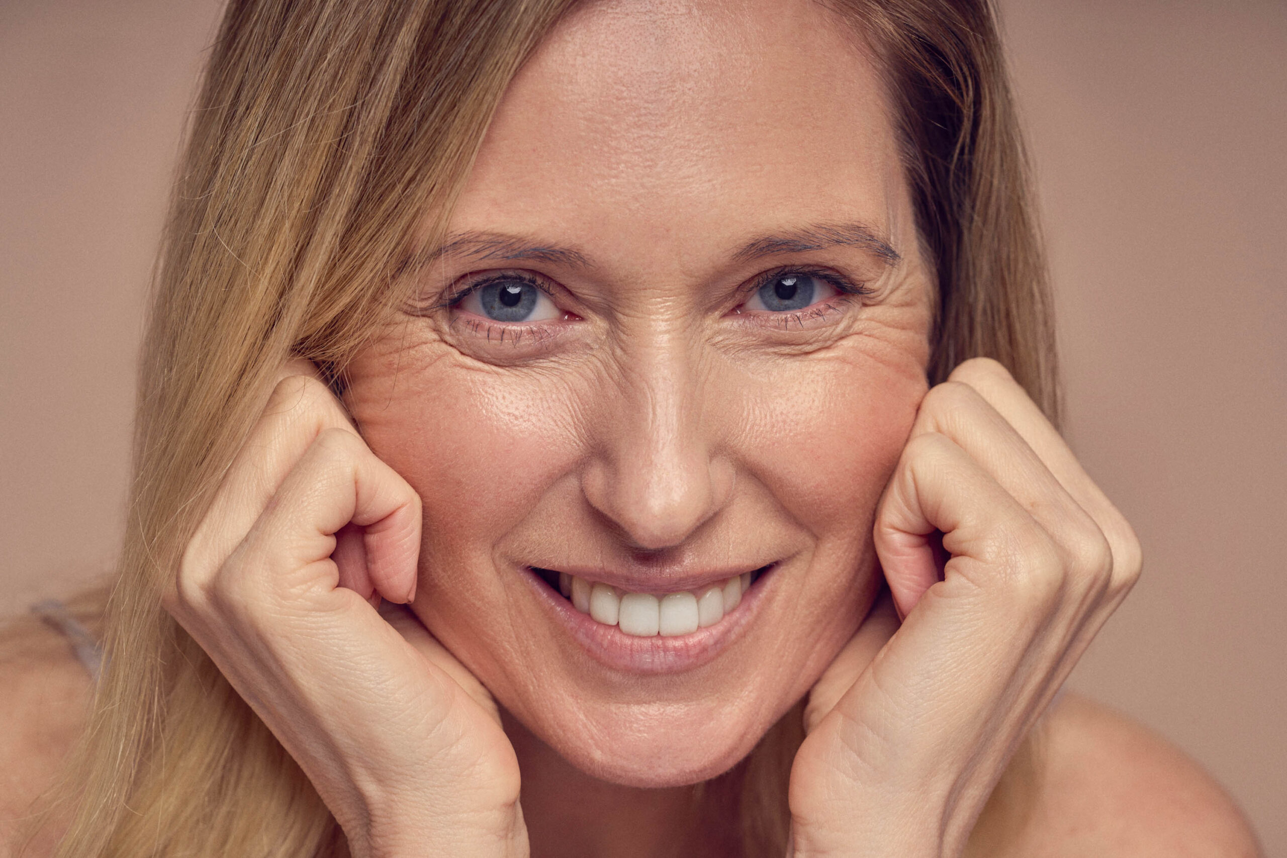 A person with long blond hair smiling, resting their chin on their hands, looking directly at the camera against a neutral background.