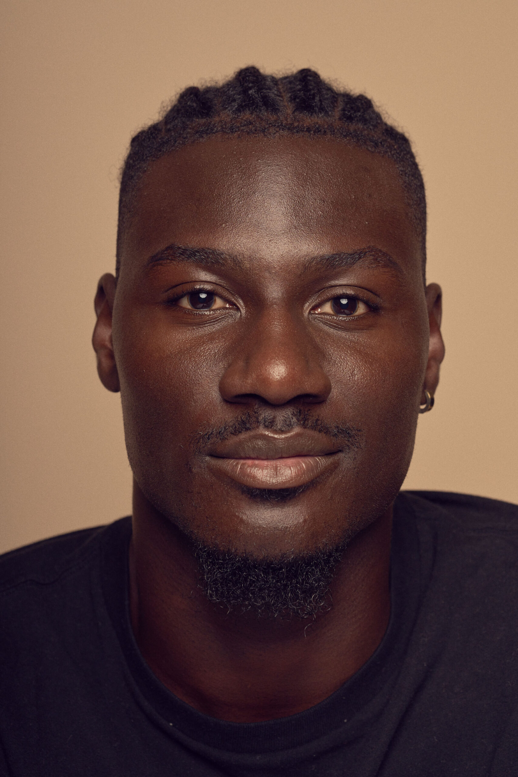 Portrait of a person with braided hair, wearing a dark shirt, set against a neutral background. They have a calm expression and are looking directly at the camera.