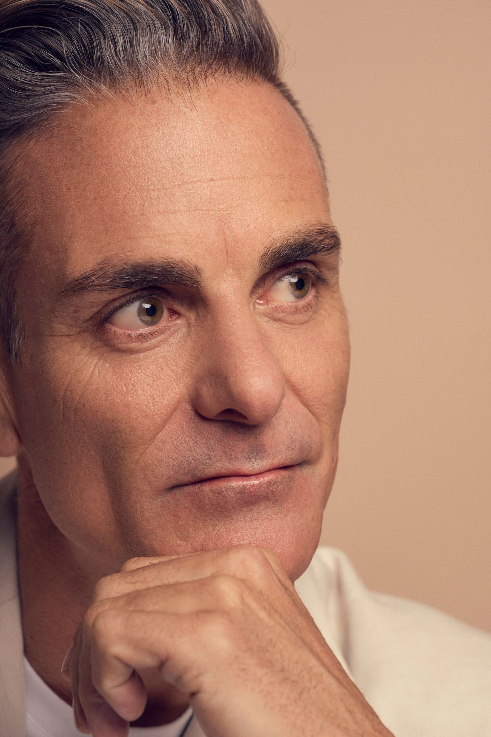 A middle-aged man with grey hair and a contemplative expression rests his chin on his hand. He wears a light-colored blazer against a neutral background.