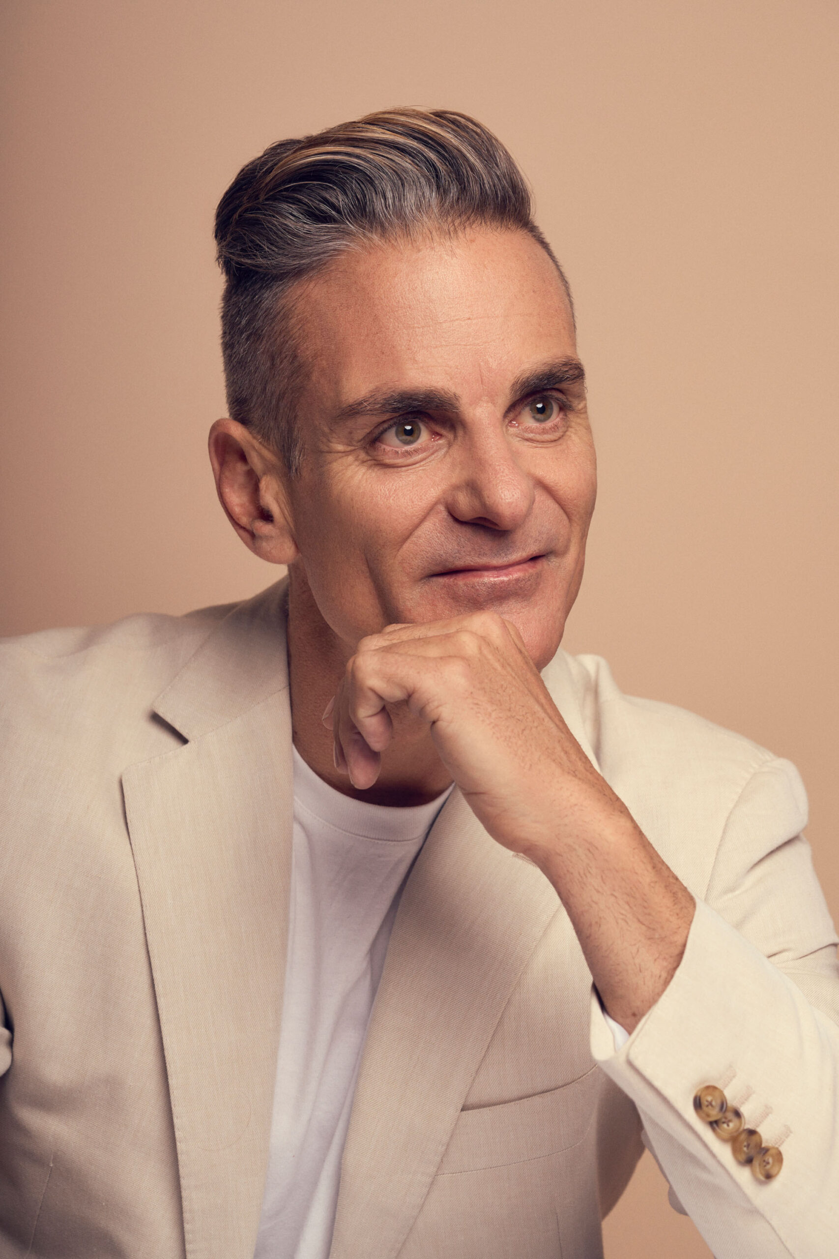 A mature man with short, styled gray hair and a slight smile rests his chin on his hand. He is wearing a beige blazer over a white shirt, set against a light brown background.