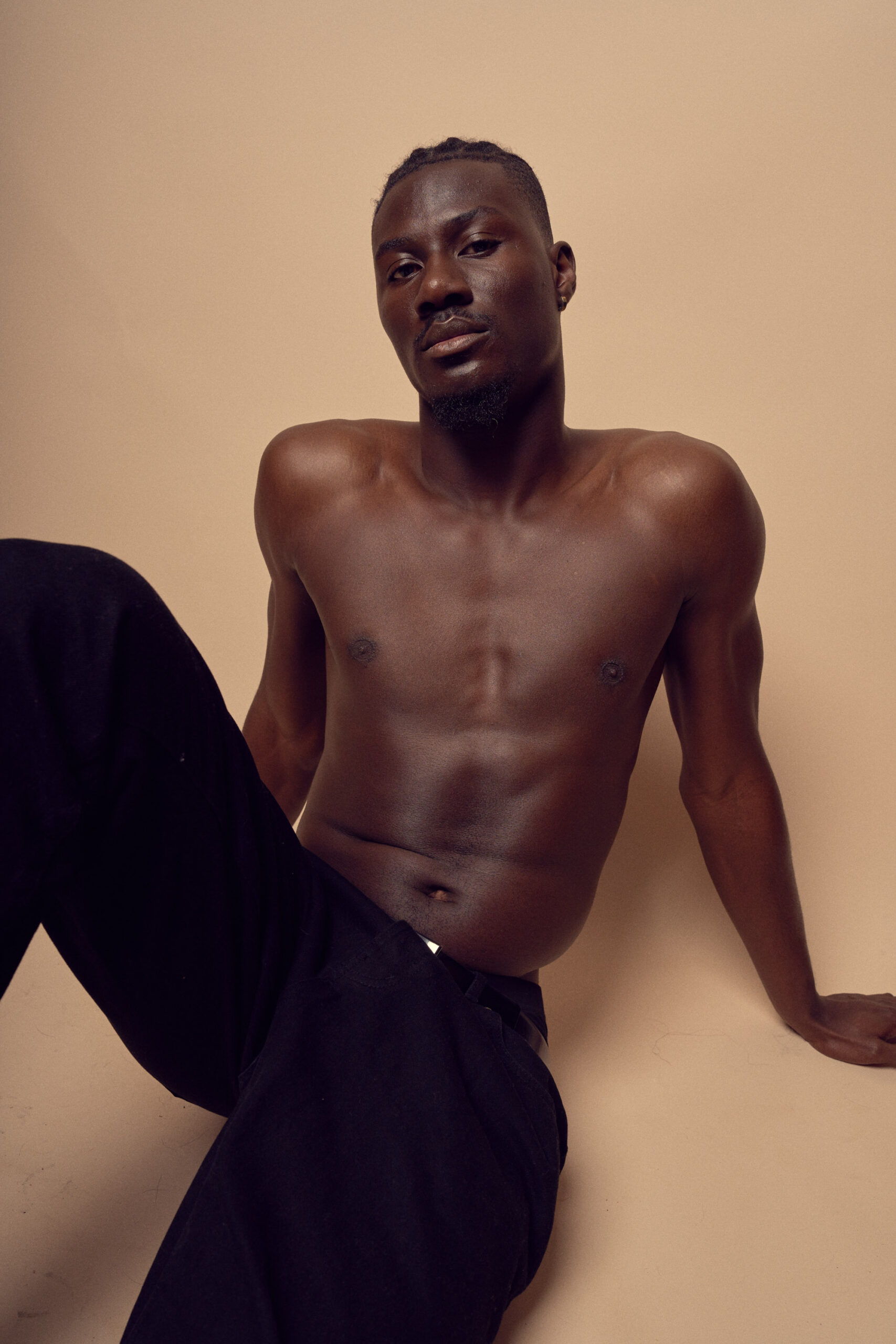 A shirtless man with short hair sits against a neutral background. He is wearing dark pants and has a relaxed expression, with one arm resting on the ground for support. The lighting casts soft shadows, emphasizing his features.