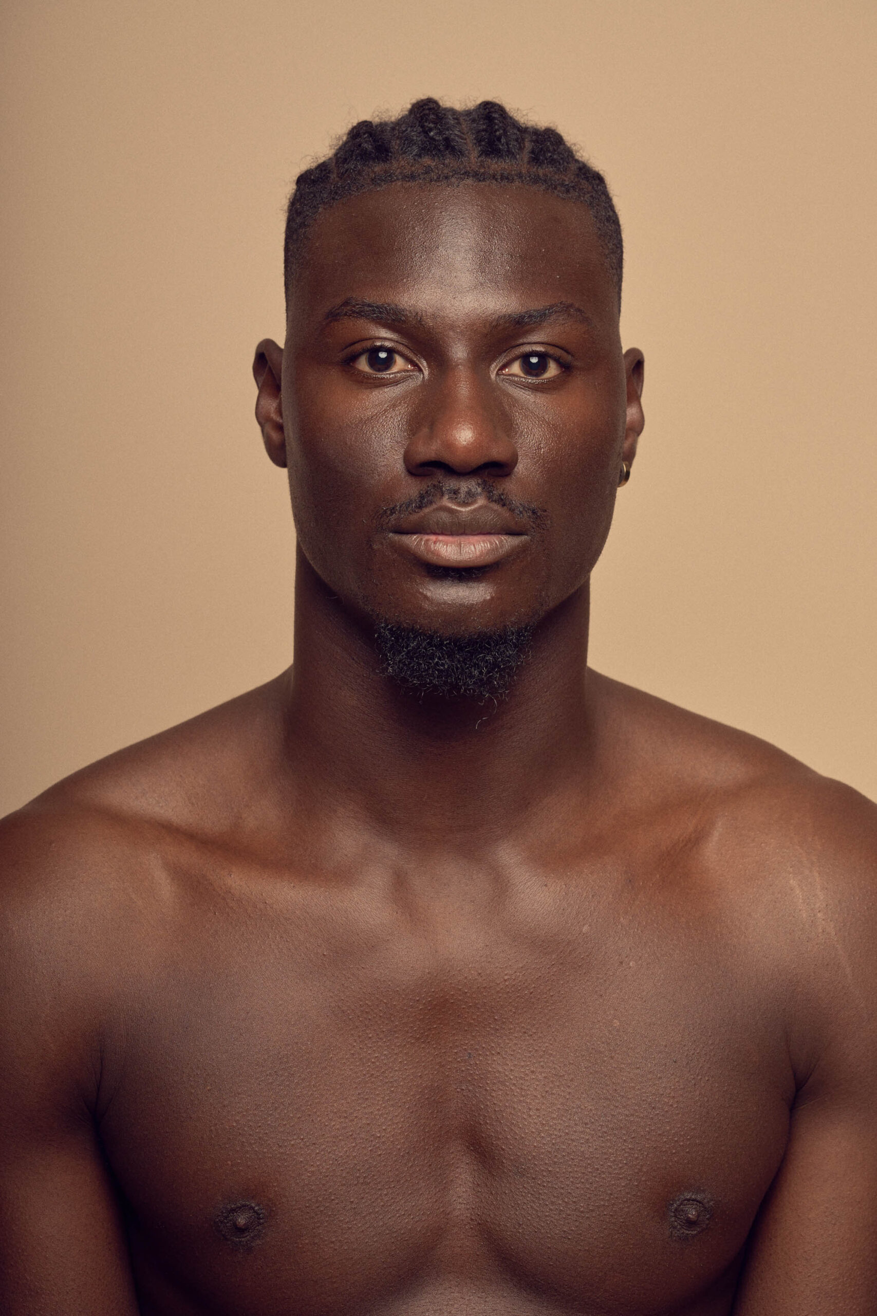 Man with short braids and earrings facing forward against a solid beige background. He is shirtless, with a calm and confident expression.