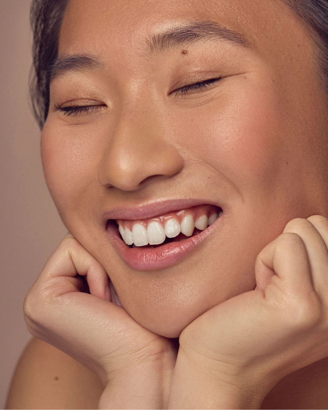 A person with closed eyes smiling joyfully, resting their chin on their hands. The background is neutral, emphasizing their radiant and expressive face.