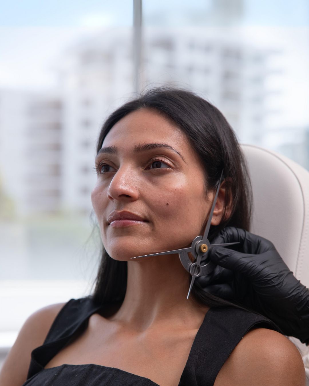 A person with long dark hair is sitting and gazing forward while a gloved hand is holding measuring calipers near their face. The background shows blurred buildings and a bright window.