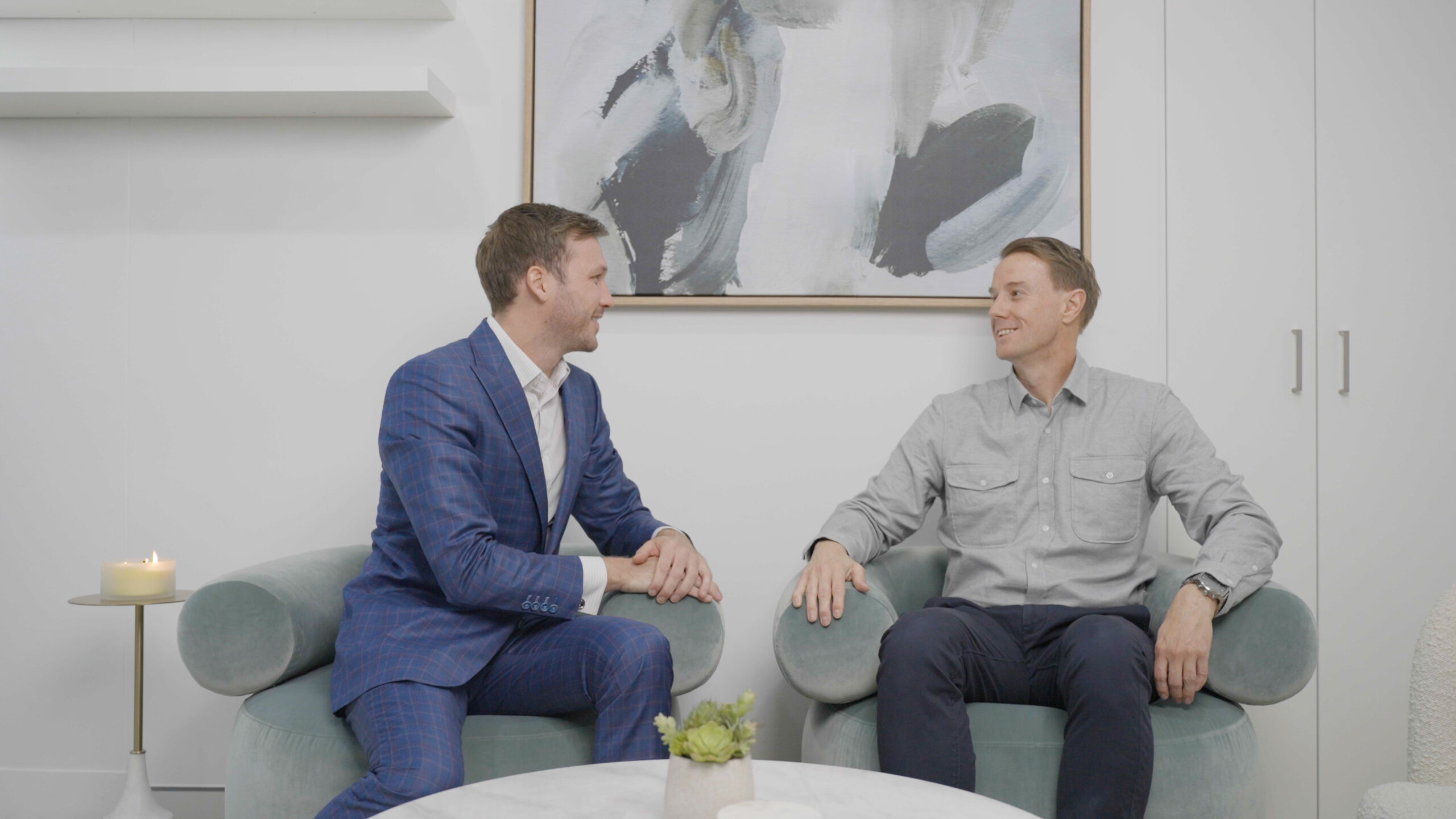 Two men sit on light green chairs in a modern office, engaged in conversation. One wears a blue suit, the other a gray shirt. A round table with a plant is in front of them, and abstract art hangs on the white wall behind.