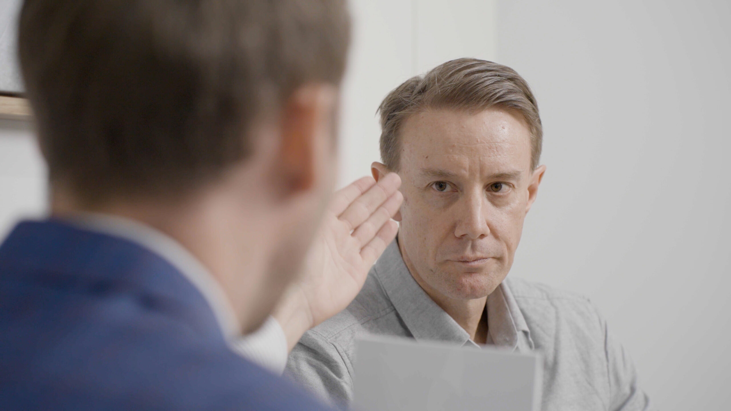 Two men are engaged in a conversation. The man on the left is gesturing with his hand, while the man on the right, wearing a light gray shirt, attentively listens and maintains eye contact. The background is a plain indoor setting.