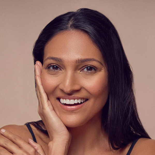 A woman with long dark hair smiles warmly at the camera. She has her hand gently touching her cheek and an arm wrapped around herself. She is wearing a sleeveless top, and the background is a soft beige tone.