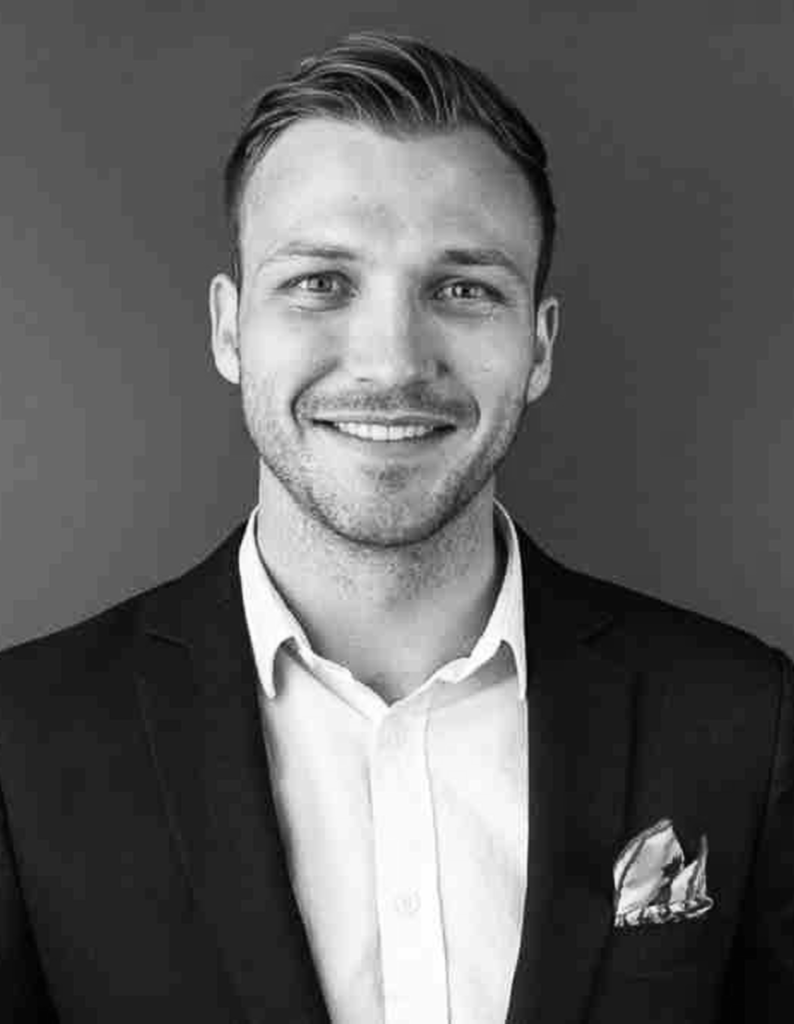 Black and white portrait of a smiling man with short hair, wearing a suit jacket, white shirt, and pocket square. He stands in front of a plain background, looking directly at the camera.