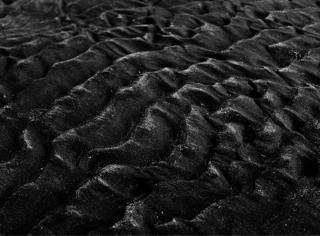 Close-up of a textured black surface resembling ripples or waves, possibly sand or fabric. The lighting creates shadows and highlights, enhancing the depth and contours of the surface.