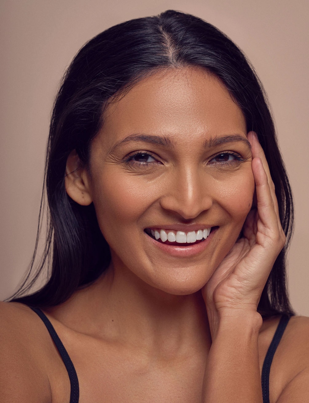 A woman with long dark hair smiles warmly, touching her face with one hand. She is wearing a black sleeveless top and is set against a neutral beige background.