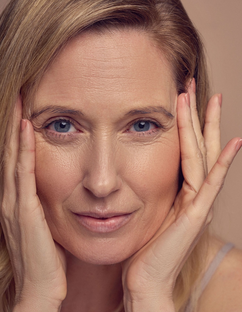 A close-up of a middle-aged woman with light skin and blue eyes, gently touching her face with both hands and looking directly at the camera with a soft expression.