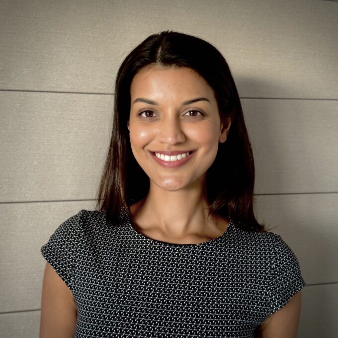 A woman with straight dark hair, wearing a short-sleeved black and white patterned top, smiles at the camera while standing in front of a light-colored, textured wall with horizontal lines.